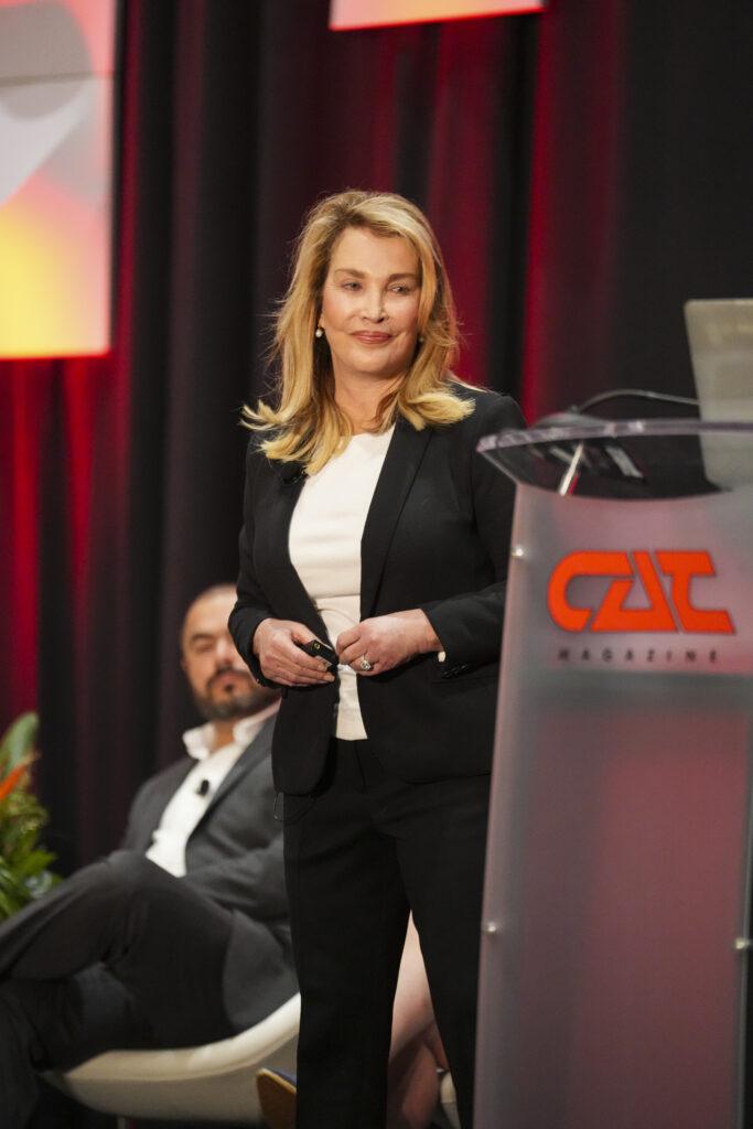 A woman in a black suit stands at a podium labeled "CAT Magazine," with a seated man in the background.