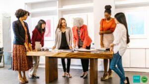 A group of six women stand around a wooden table, engaged in discussion over documents in a modern office setting.