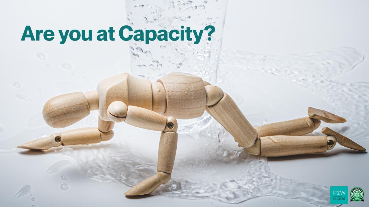 A wooden mannequin on hands and knees is surrounded by spilled water, with the text "Are you at Capacity?" above.